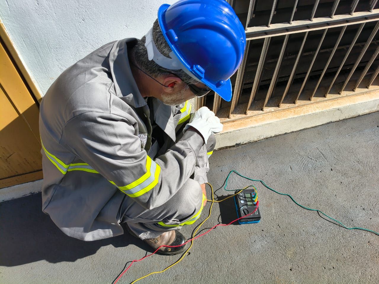 Técnico da IC Para Raios com EPI completo — capacete, óculos, protetor auricular e luvas — operando terrômetro em medição de aterramento